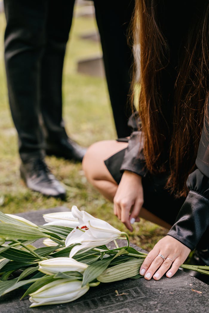services-01 A woman kneels by a grave adorned with white lilies, symbolizing loss and remembrance.